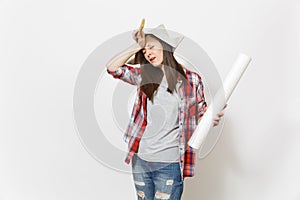 Young exhausted woman in newspaper hat holding wallpaper roll and paint brush and keeping hand near face isolated on