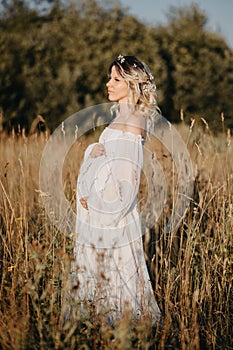 A young expectant mother in the third trimester of pregnancy in a white dress hugs her stomach against the backdrop of a
