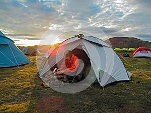 Young experienced hiker active man is cooking dinner outdoors next to the tent in a wild nature campsite at sunset