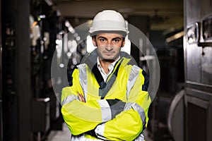 Young factory engineer or worker wearing safety vest and hard hat crossing arms at electrical control room. Industry and