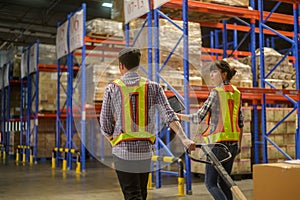 Young factory workers pulling a pallet truck between shelves full of packed boxes in warehouse, machinery and Logistics concept