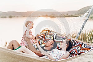 Young family with baby on the beach, smiling father and mother holding cute little girl while lying in a hammock, hipster family