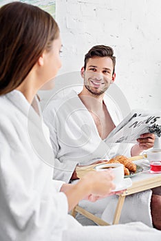 young family in bathrobes having breakfast and reading newspaper