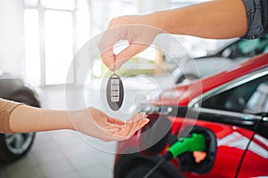 Young family buying first electric car in the showroom. Close-up of male hand giving car key to female hand on battery