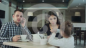 Young family in cafe. Father make a phone call while mother and daughter talking. Mother looking at him dissatisfied.