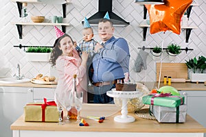 Young family celebrating first birthday of their little baby boy with a cake, present boxes, and air balloons. Birthday