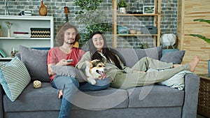 Young family and corgi dog watching TV on couch at home laughing relaxing