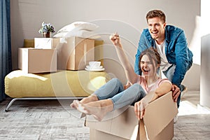 Young family couple bought or rented their first small apartment. Cheerful woman scream sitting in box. Guy move her