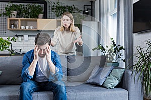 Young family, couple at home, man and woman quarrel, conflict, woman shouting and waving hands. Man sitting on sofa, holding head