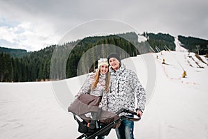 Young family couple near baby stroller on snow in Carpathian mountains. On background of forest and ski slopes with lifts. Close