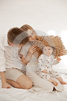 Young family couple sit on bed and dress up little baby wicker hat on white background. Home family photo of mom, dad