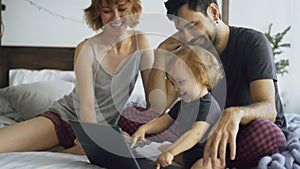 Young family with cute little girl playing with daughter while she typing on laptop computer sitting in bed at home