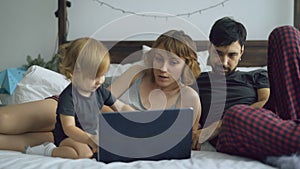 Young family with cute little girl playing with daughter while she typing on laptop computer sitting in bed at home