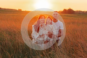 Young family with daughter sits on meadow among grass at sunset