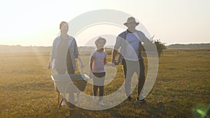 Young family having fun outdoors in their farm. Gardener woman pushing wheelbarrow with vegetables at sunset. Family