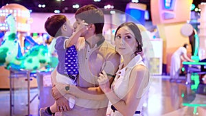 Young family with little child in amusement park, father is holding son on hands, his wife is standing near