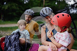 Young family with little children resting after bike ride, sitting on grass in park in summer.