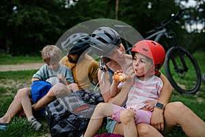 Young family with little children resting after bike ride, sitting on grass in park in summer.