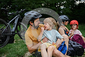Young family with little children resting after bike ride, sitting on grass in park in summer.