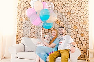 Young family man, woman and baby girl sitting on couch indoors with wooden decorative wall and multi-colored balloons with helium