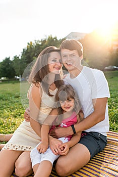Young family man woman child daughter 5-7 years old, summer park, sitting on blanket, relaxing, posing in nature