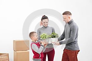 Young family, man woman and child son in new apartments. They are holding green potted plants. Boxes with cargo on a