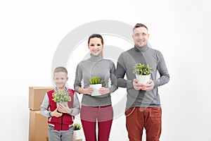 Young family, man woman and child son in new apartments. They are holding green potted plants. Boxes with cargo on a