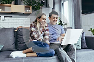 Young family man and woman having fun and happily sitting at home using laptop and smiling, multiracial couple