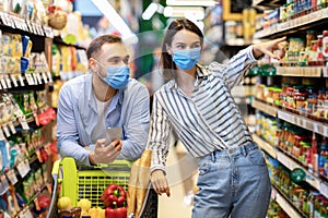 Young family in masks with phone shopping in hypermarket