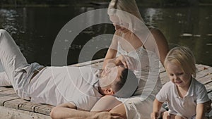 Young family mom father and son are sitting on the pier near the river on a sunny day. A man lies on a woman lap