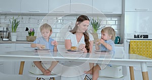Young family mom and two sons sitting in the kitchen at the white table eating burgers together and having fun. Kids