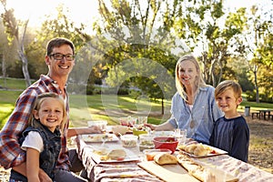 Young family at a picnic in a table in a park look to camera