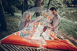 Young family playing with son on a hammock in park on camping trip, hipster mother putting a vintage hat on a baby boy near