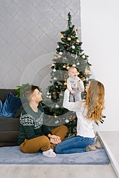 A young family sits on the floor in the living room near the Christmas tree, mom lifted up the baby in her arms, they are happy