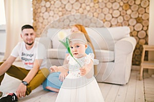 Young family sits on the floor near the couch,small daughter of a blonde one year old is learning to walk in a white dress against