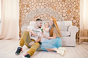 Young family sits on the floor near the couch,small daughter of a blonde one year old is learning to walk in a white dress against