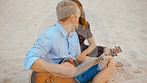 Young family is sitting on sand on beach playing guitar and singing in summer
