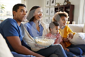 Young family sitting together on the sofa in their living room watching TV and eating popcorn, side view photo