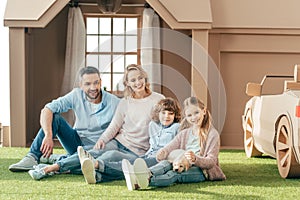 young family sitting on yard of cardboard house