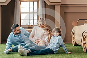 young family sitting on yard of cardboard house