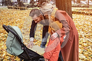 A young family is walking in an autumn park with a son and a newborn baby in a stroller. Family outdoors in a golden