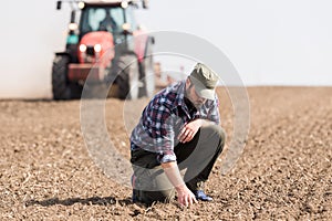 Young farmer examing dirt while tractor is plowing field