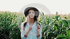 Young farmer girl in a hat, on a corn field, goes through the tall corn stalks in the sun. Cultivation of agricultural