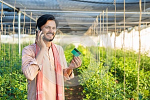 young farmer at greenhouse talking on mobile phone call by holding green credit card - conept of online payment