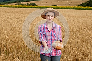 Young farmer holding dollars and bread in golden wheat field. Profit from agriculture during harvesting season in the