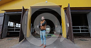 Young farmer owner in shirt and orange vest looking at camera in background of dairy farm. Cowshed barn interior, stall