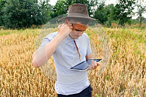 Young farmer. Portrait of farmer seating in gold wheat field and scrolling on tablet. Young man wearing cowboy hat in