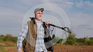 A young farmer with a shovel on his shoulder walks along fields in the countryside