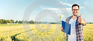 Young farmer standing in wheat field with clipboard, looking directly at camera and showing thumbs up