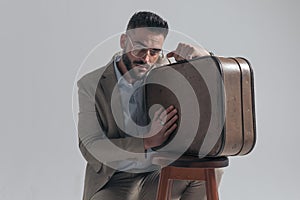 Young fashion model with beard posing and holding suitcase on wooden chair